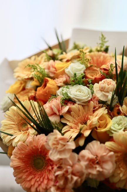 A vibrant floral arrangement featuring a mix of fresh flowers including peach and cream-colored gerbera daisies, soft pink roses, white lisianthus, and yellow-orange spray roses, accented with lush gr