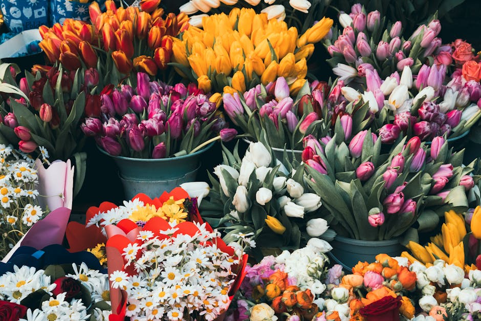 A vibrant display of freshly cut tulips in various colors including purple, white, pink, yellow, and orange arranged in metal buckets. Surrounding the main arrangement are bouquets of mixed flowers su