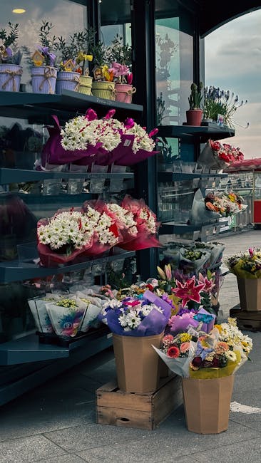 An outdoor flower display at Wapping Florist showcases a variety of floral arrangements and bouquets on metal shelves and wooden crates. The flowers include vibrant pink, white, purple, and yellow blo