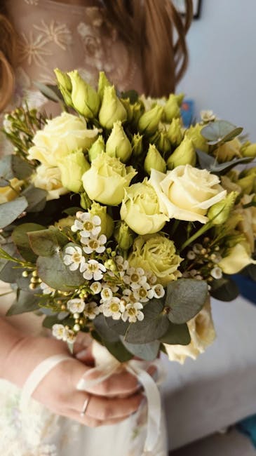 A close-up of a bridal bouquet held by a person wearing a lace wedding dress, featuring cream and pale yellow roses, white waxflowers, and eucalyptus leaves. The arrangement is tightly clustered, crea