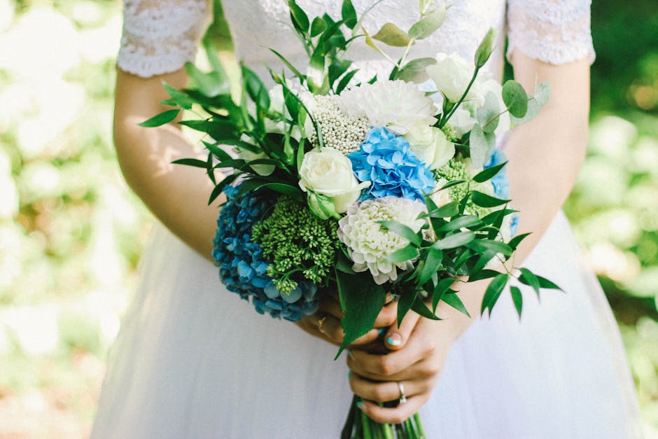 A bride in a white lace dress holding a bouquet of fresh flowers outdoors. The bouquet features blue and white hydrangeas, white roses, and green foliage, arranged in a loose, natural style. The flowe